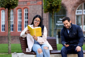 Two postgraduate students sitting on a bench, one reading a book.