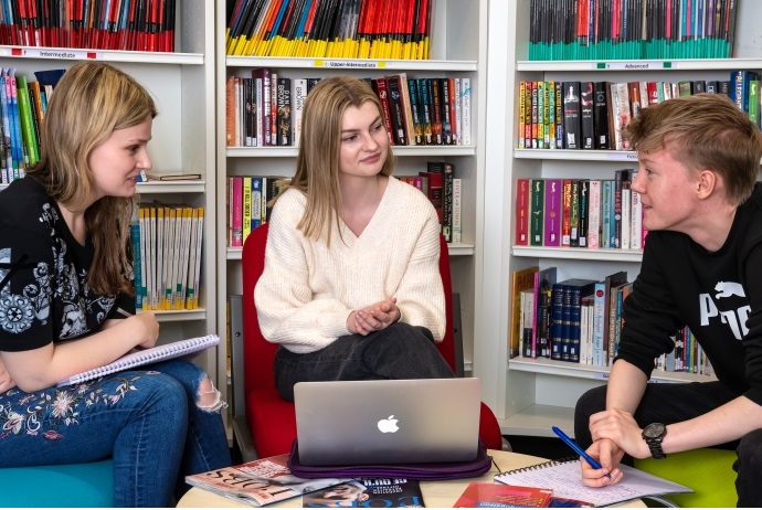 Three students with laptops sit together in a library.