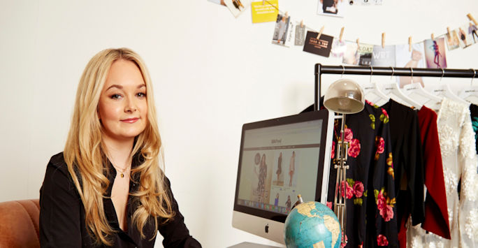 Emma Watkinson sitting at her desk with her computer and a rail of clothing.