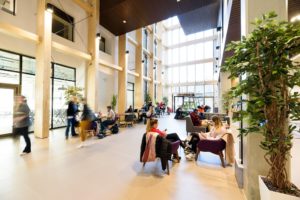 Students sit, scattered around the different seating areas of the bright and airy foyer of the School and Social Justice Building.